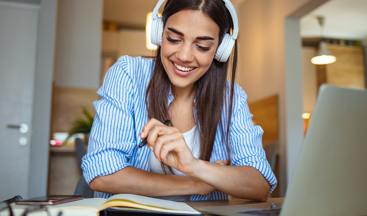 A young brown woman smiling, wearing headphones while reading a book with a laptop on her side.
