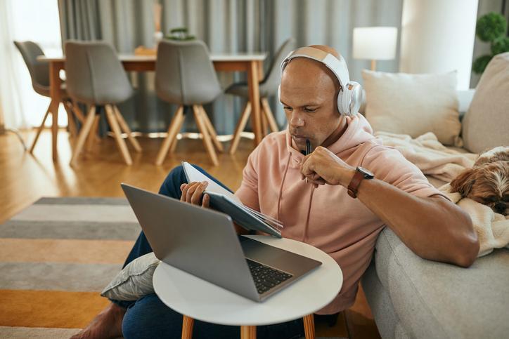 A man wearing headphones and holding a notebook looks at a laptop computer.